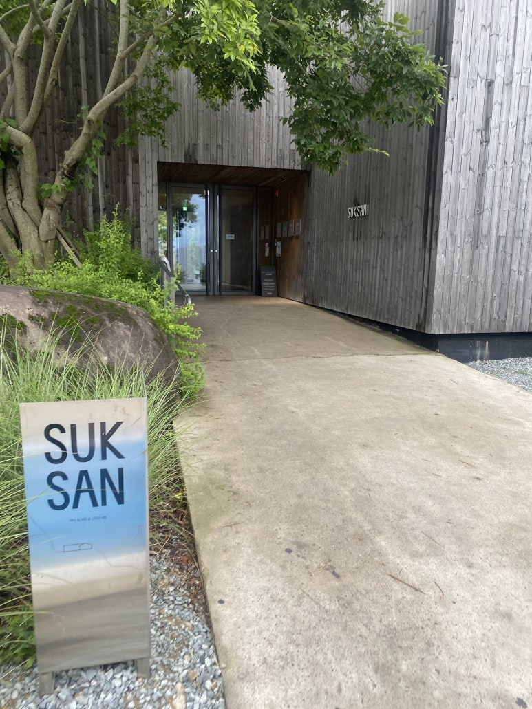 나무 외벽과 SUKSAN 표지가 보이는 입구